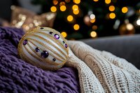 Gold striped ornament with purple gemstones resting on cozy knit fabrics, warm bokeh lights in background, festive holiday decoration for seasonal celebrations.
