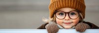 Young child wearing orange knit beanie and round glasses smiling warmly while resting chin on hands in cozy brown sweater against soft neutral background.