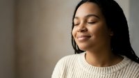Young African American woman with closed eyes and peaceful expression wearing cream sweater in soft natural light for wellness and mindfulness concepts.