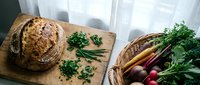 Artisan sourdough bread with fresh herbs on cutting board next to wicker basket filled with organic root vegetables including carrots, beets, radishes.