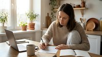 Young woman working from home office writing notes while using laptop computer in cozy kitchen workspace with plants and natural lighting.