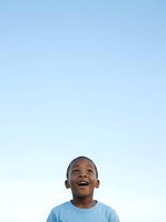 Young boy, African descent, wearing light blue t-shirt, looking upward with joyful expression against clear sky background. Suitable for childhood, happiness, optimism, and aspirational themes.