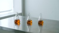 Amber liquid in three glass bottles arranged on reflective surface with window light, suitable for beverage, alcohol, or product comparison concepts.