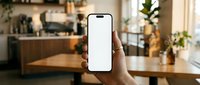Hand holding a smartphone with blank white screen mockup in a cozy cafe interior, perfect for app and UI presentation designs.