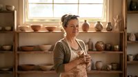 Middle-aged Caucasian female potter in apron standing in ceramic workshop studio surrounded by handmade clay pottery bowls and vases on wooden shelves.