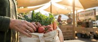 Mature man holding fresh vegetables in paper bag at farmers market with produce stand in background.