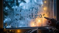 Hand writing Merry Christmas on a foggy winter window with glowing lights, pine branches, and a cozy mug, evoking holiday warmth.