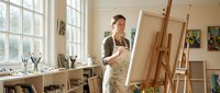Young female artist painting on canvas in bright art studio with easels, brushes and creative workspace for artistic expression and professional development.