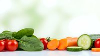Fresh organic vegetables including cherry tomatoes, spinach leaves, sliced carrots and cucumber arranged on white surface for healthy cooking and nutrition.