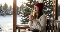 Young Caucasian woman in red knit hat enjoying hot drink on wooden cabin porch during winter morning with snow-covered forest background.