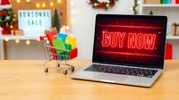 Laptop with glowing red Buy Now screen beside a mini shopping cart with colorful bags, Christmas decor and seasonal sale sign in background.