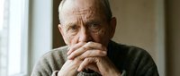 Thoughtful elderly Caucasian man with hands clasped near face in contemplative pose by window. Senior male reflecting indoors with natural lighting.