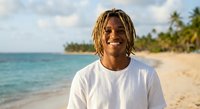 Young African American man with dreadlocks smiling on tropical beach with palm trees and turquoise ocean water during golden hour vacation lifestyle.