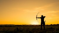 Silhouette of archer drawing bow at golden sunset in open field, suitable for sports, archery training, outdoor recreation, and motivational content.