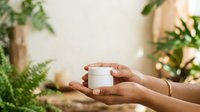 Hands holding white cosmetic jar with green plants in background, skincare product presentation for beauty and wellness marketing.