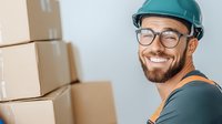 Cheerful Caucasian male warehouse worker with beard and glasses wearing hardhat smiles while organizing cardboard boxes for shipping and delivery service.