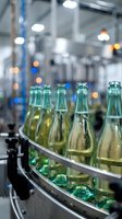 Filled glass bottles with carbonated beverage moving on production line conveyor belt in modern factory facility with blurred industrial background and overhead lighting.