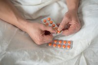 Senior man hands holding orange medication pills blister packs on white bedding for healthcare and treatment management.