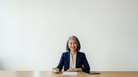 Mature woman with gray hair, East Asian ethnicity, wearing navy blazer and cream blouse, seated at wooden desk with coffee cup, professional business portrait for corporate and executive use.