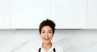 Young woman with short, curly dark hair and olive complexion wearing black and white chef's uniform, smiling enthusiastically in modern kitchen with marble countertop and white cabinetry, suitable for culinary, hospitality, and professional cooking content.