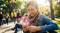 Happy young Caucasian woman using smartphone in sunny park with warm golden light and lens flares creating joyful atmosphere for lifestyle and technology content.