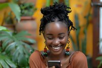 Young African woman with natural hair and decorative earrings smiling while looking at mobile phone, surrounded by green tropical plants in warm outdoor setting.