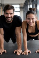 Caucasian athletic couple doing plank exercise together at gym, smiling at camera. Young man and woman in black sportswear working out on fitness mat.