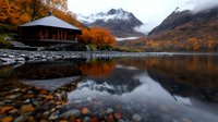 Tranquil mountain lake with wooden pavilion surrounded by vibrant autumn foliage, snow-capped peaks and misty clouds reflecting in calm water surface.