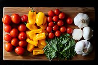 Fresh organic ingredients on wooden cutting board: ripe cherry tomatoes, sliced yellow bell peppers, fresh basil leaves and whole garlic bulbs arranged for cooking.