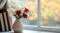 Fresh white and red flowers in modern ceramic vase on windowsill with books, soft natural daylight creating cozy home atmosphere with blurred autumn background.