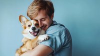 Young Caucasian man in blue t-shirt hugging and smiling with happy Welsh Corgi dog against blue background, showing pet love and companionship.