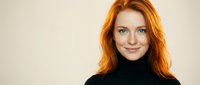 Young Caucasian woman with long red hair wearing black turtleneck sweater smiling at camera against neutral beige background for portrait photography.