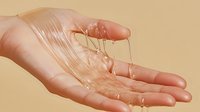 Dripping honey-like golden substance flowing from palm on beige background, viscous liquid creating thin glossy strands showing texture and consistency.