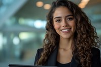 Young Hispanic businesswoman with curly brown hair and bright smile wearing black blazer in modern office setting, holding digital tablet. Professional corporate portrait.