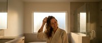 Young woman with long wavy hair in white bathrobe touching her hair in modern luxury bathroom with warm lighting and minimalist design.