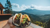Mexican street tacos with guacamole and meat held against scenic mountain landscape view from wooden deck, forest and snow capped peaks in background on sunny day.