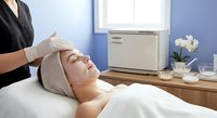 Young Asian woman receiving facial treatment at spa, esthetician applying mask in serene blue wellness room with towel warmer.