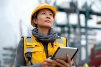 Female engineer in yellow hard hat and safety vest using digital tablet at industrial port, looking up at shipping infrastructure.