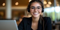 Young Hispanic businesswoman wearing headphones and glasses smiles while working on laptop in modern office space with bokeh background lighting.