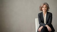 Middle-aged woman with wavy gray hair, Caucasian ethnicity, wearing professional black blazer and white blouse, seated on white chair against neutral gray background for corporate and business use.
