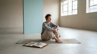 A young man with brown hair and light skin sits cross-legged on a yoga mat in a bright, minimalist studio space with large windows, wearing casual athletic wear, ideal for wellness, fitness, and lifestyle content.