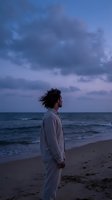 Young man with curly hair in white clothing standing on beach at twilight looking at ocean waves under dramatic purple sky for wellness and mindfulness concepts.