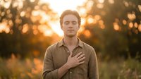 Young Caucasian man with reddish-brown hair wearing a tan button-up shirt, hand on chest, standing outdoors during golden hour sunset with blurred trees and warm bokeh background.