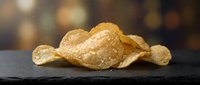 Golden crispy potato chips with sea salt on dark slate surface against warm bokeh background for snack food packaging and culinary marketing materials.