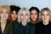Diverse group of young women with different hair colors and skin tones wearing dark clothing pose together in studio portrait showing beauty and inclusivity.