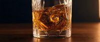 Premium whiskey in crystal cut glass with ice cube creating elegant swirl pattern on wooden surface against dark background for luxury beverage advertising.