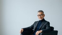 Mature male professional with gray hair and glasses wearing dark blazer and navy shirt, seated in relaxed pose against neutral background for corporate portraits.