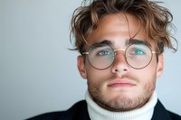 Young Caucasian male model wearing round glasses and white turtleneck sweater against light background, close up portrait showing bright blue eyes and styled brown hair.