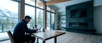 Professional businessman working on laptop at wooden desk in modern luxury home office with mountain view and contemporary fireplace design.