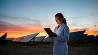 Female engineer with tablet inspecting solar panel farm at sunset. Renewable energy technology and sustainable power generation concept.